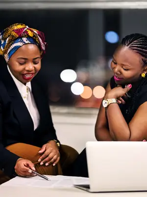 Two women review documents together, smiling, with city lights visible through a window.