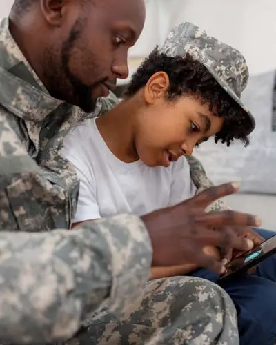 Photo of a father in miltary uniform looking at a phone with his tween son