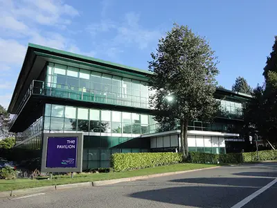 A modern glass building stands prominently, surrounded by greenery and a clear blue sky.