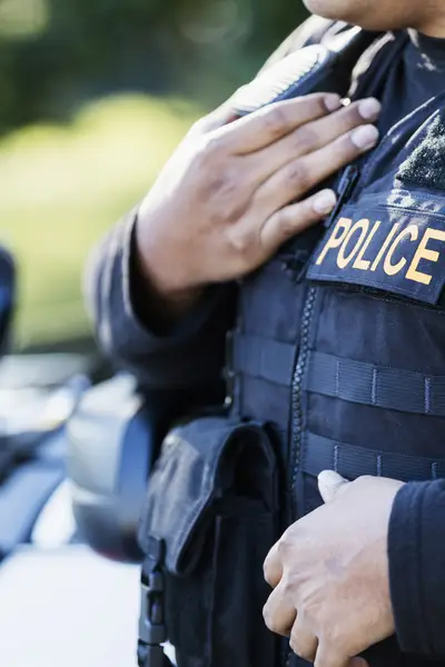 A police officer's torso is shown with a police car in the background.