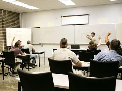 Adult students in classroom raising hands while instructor teaches.