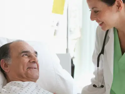 A female doctor stands next to the hospital bed of an older male patient
