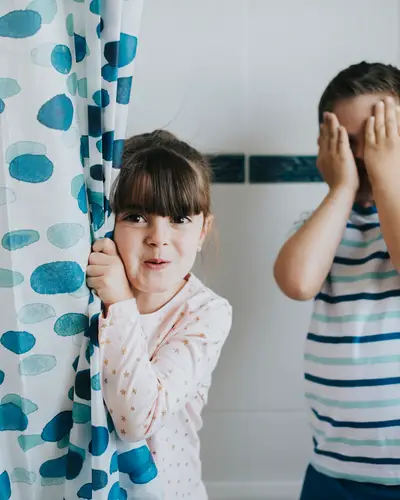 Two kids in a bathtub fully clothed and appearing around the corner of the shower curtain