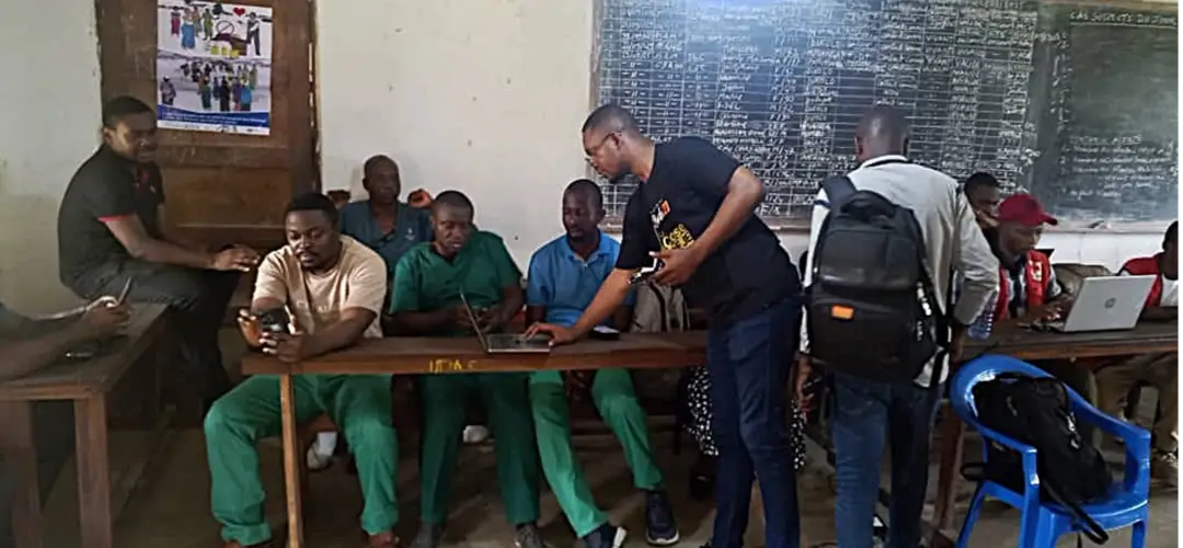 men gathered around a table. RTI employee Xavier Kalamba mentors laboratory technicians on lab results data entry.