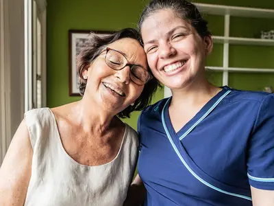 A senior woman smiles and leans on her caregiver's shoulder.