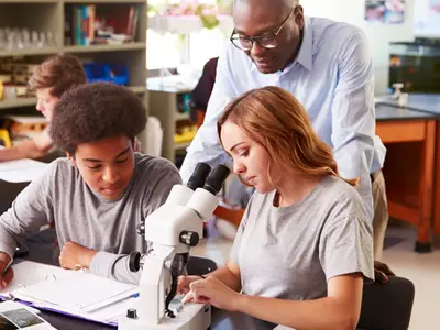 Two high school students use a microscope with help from a male science teacher.