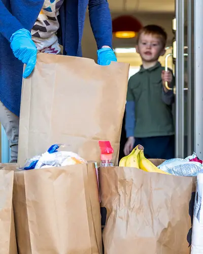 A delivery person brings bags of groceries to a family's front steps, while a boy watches from inside the door.
