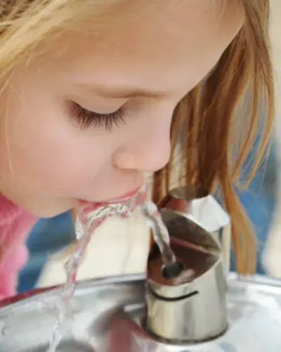 A girl drinks from a metal water fountain in a public outdoor area.