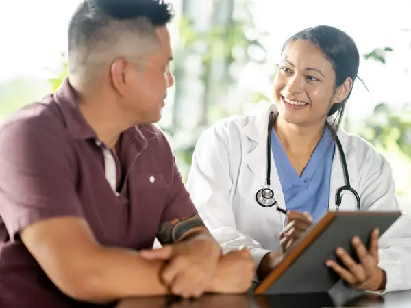 A man speaks with a female doctor who is wearing a stethoscope and holding a clipboard.