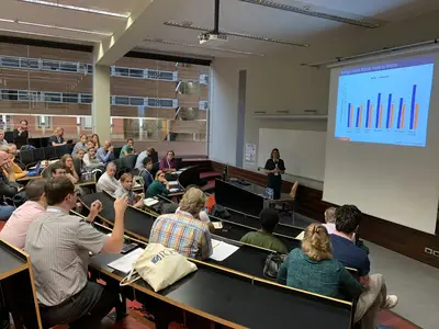 A presenter discusses data on a projector while an audience engages in a seminar setting.