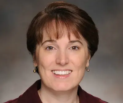 A woman smiles warmly, wearing a dark blouse, with a neutral background.