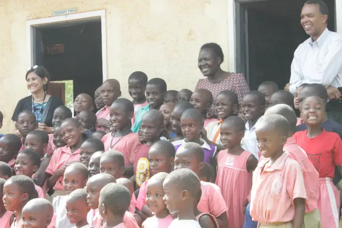 A group of children and two adults smile outside a beige building in daylight.