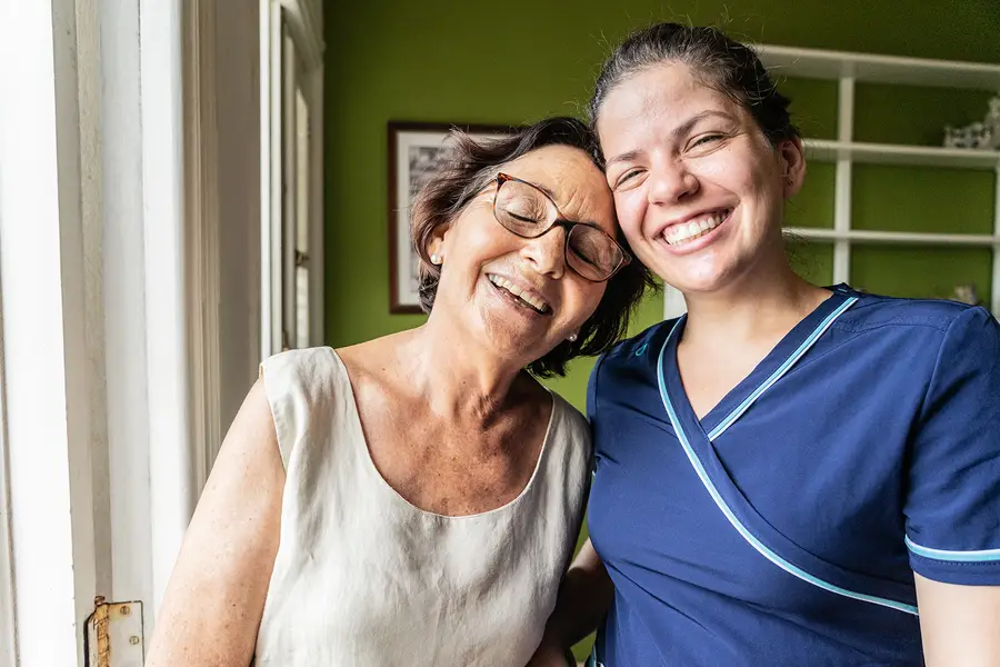 A senior woman smiles and leans on her caregiver's shoulder.