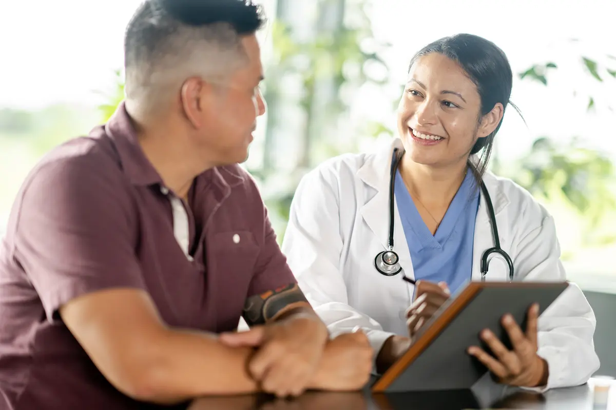 A man speaks with a female doctor who is wearing a stethoscope and holding a clipboard.