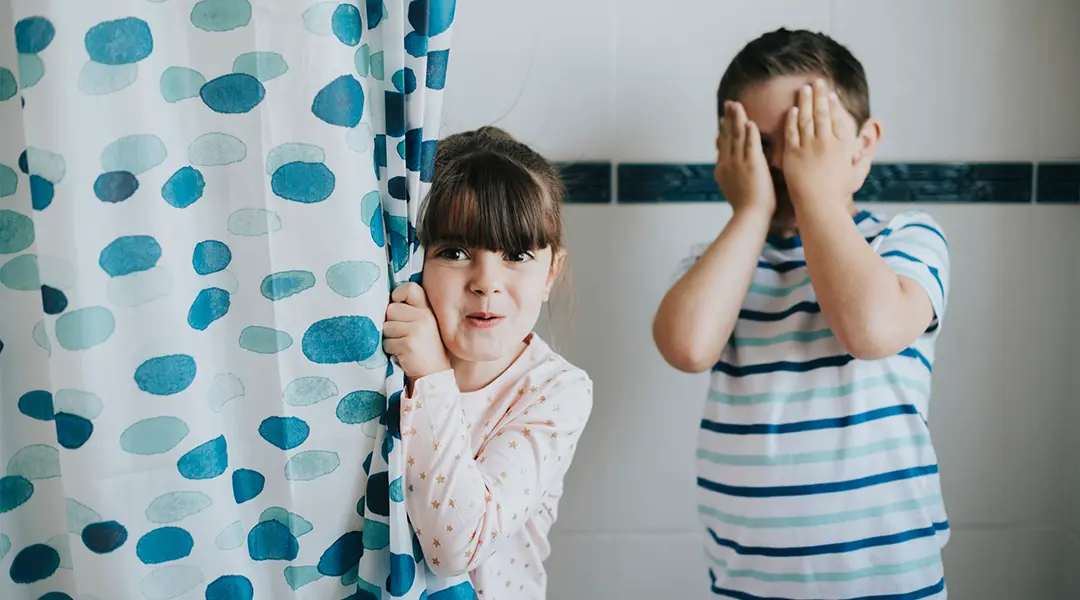 Two kids a bathtub fully clothed and appearing around the corner of the shower curtain