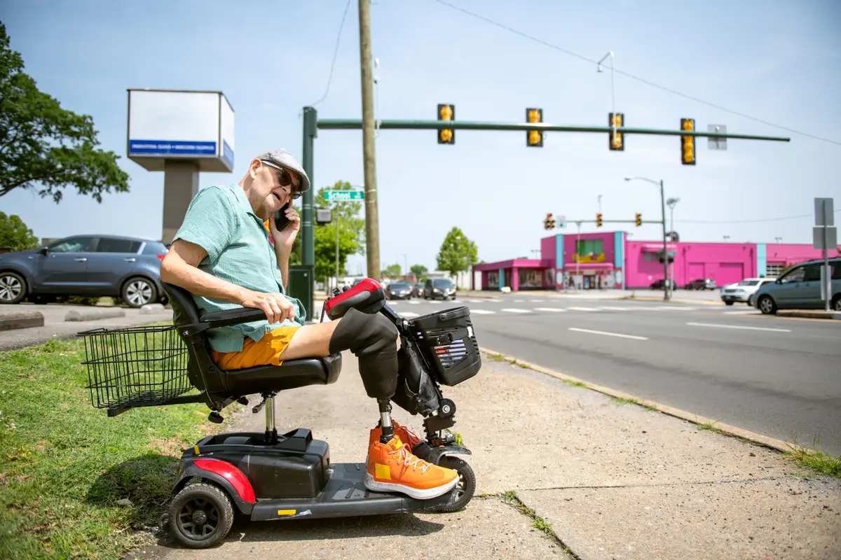 A senior man with two prosthetic legs sits in a wheelchair and calls for a ride from a sidewalk next to a major road.
