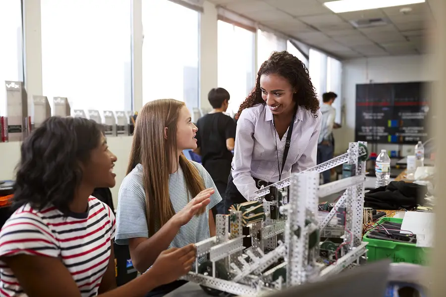 Three girls engage in conversation about a robotic project in a bright classroom.