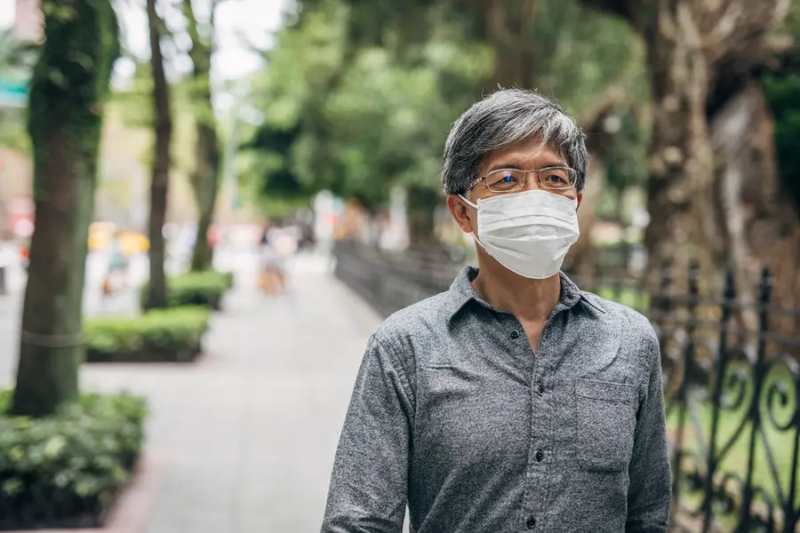 An Asian senior man wears a face mask and walks along a tree-lined sidewalk.