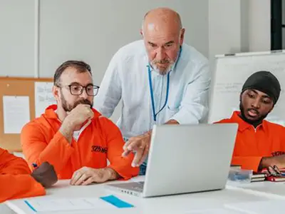 Incarcerated men looking at a laptop. An instructor pointing to the laptop's screen