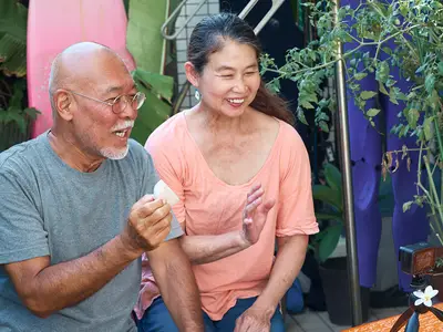 Sitting in front of a surfboard in a tropical setting, a senior Asian couple records a video for their blog.