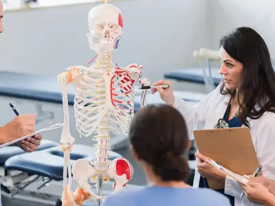 Young adult students in an anatomy class gather around an instructor and a model of a skeleton.