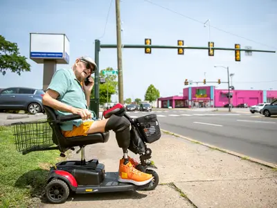 A senior man with two prosthetic legs sits in a wheelchair and calls for a ride from a sidewalk next to a major road.