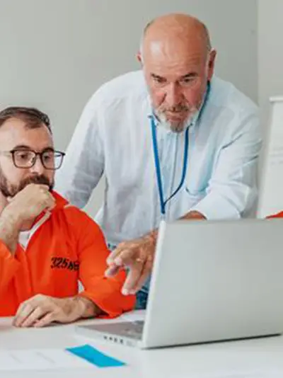 Incarcerated men looking at a laptop. An instructor pointing to the laptop's screen