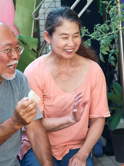 Sitting in front of a surfboard in a tropical setting, a senior Asian couple records a video for their blog.