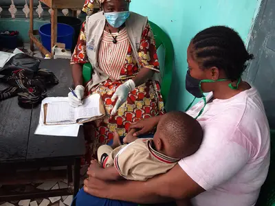 Healthcare worker records information while mother holds her child; both seated in a residential area.