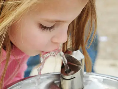 A girl drinks from a metal water fountain in a public outdoor area.