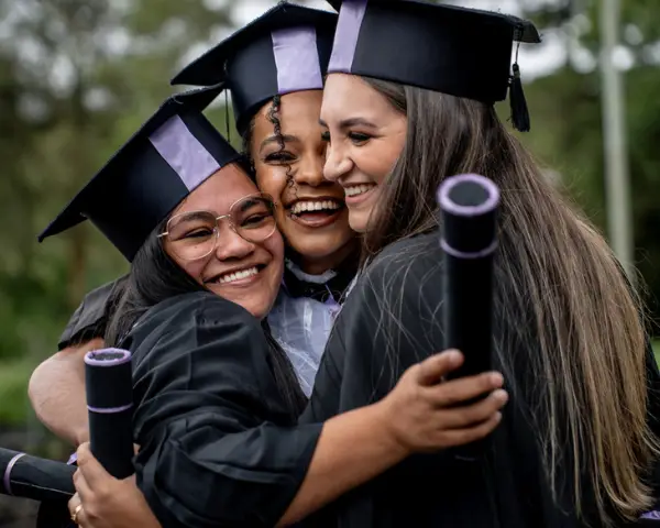 Three graduates in caps and gowns hugging and smiling while holding their diplomas.
