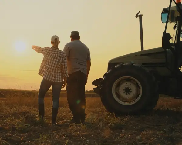 A man and woman stand next to a tractor and look out over the horizon on a farm at sunset.