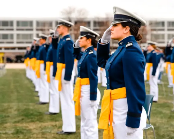 Cadets in blue uniforms and yellow sashes salute, standing in rows on a grassy field.