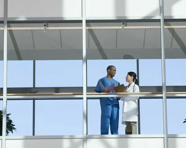 A male and female doctor talk in a glass-walled hospital walkway.