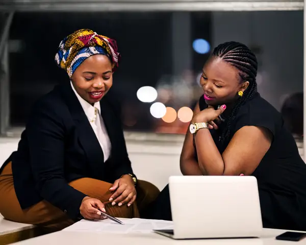 Two women review documents together, smiling, with city lights visible through a window.