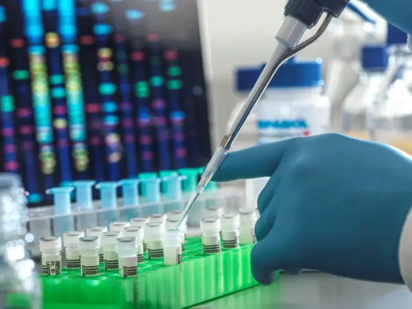 Close up photo of a scientist pipetting liquid into tubes in front of a colorful computer screen