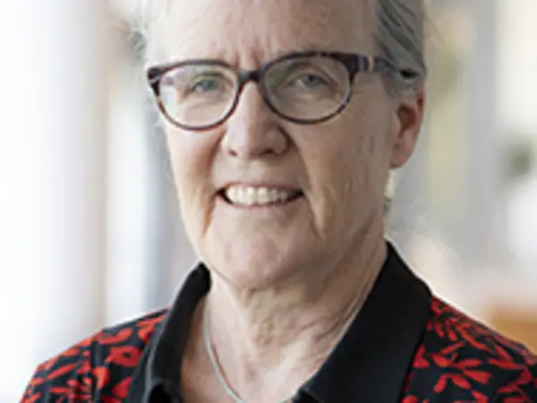 A smiling elderly woman wears a patterned shirt while standing in a bright indoor space.