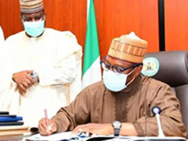 Man wearing a mask signing documents at a desk; another man stands beside him watching.