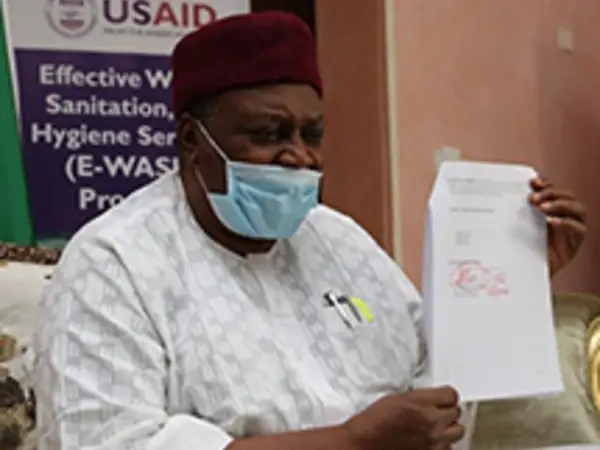 Man wearing a mask holds document in hand, seated beside a USAID sanitation program banner.