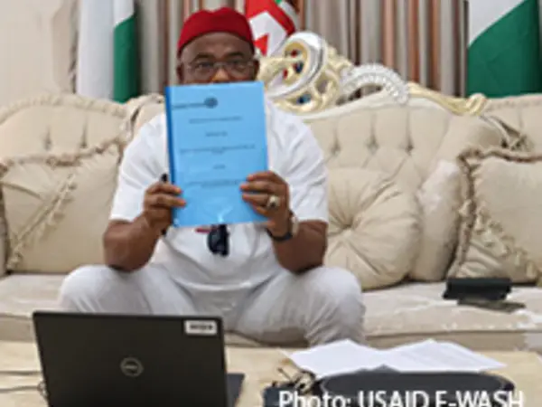 A man holds a blue document while sitting on a beige couch in an office setting.