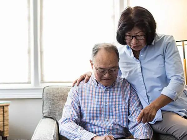 An elderly man sits on a couch while a woman assists him with a tablet.