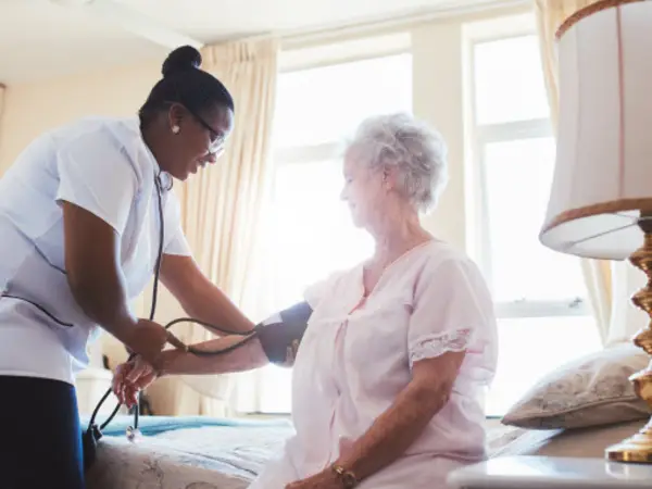 Nurse taking elderly woman's blood pressure in a sunlit bedroom with bed and lamp.