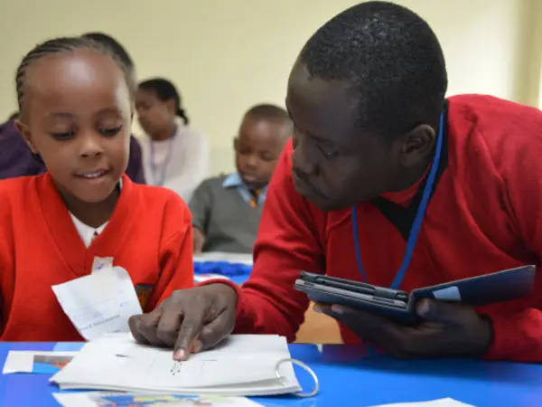 Teacher instructing a student seated at a desk in a busy classroom setting.