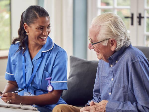 Caregiver smiles at elderly man, sitting and chatting together in a cozy living room.