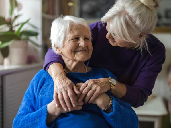 Elderly woman embraces younger woman in cozy, well-lit home near bookshelf and potted plant.