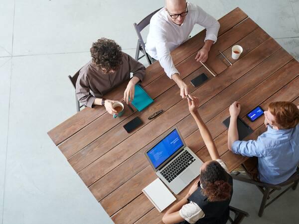 Four people at a wooden table talking, using laptops, notebooks, and drinking coffee.